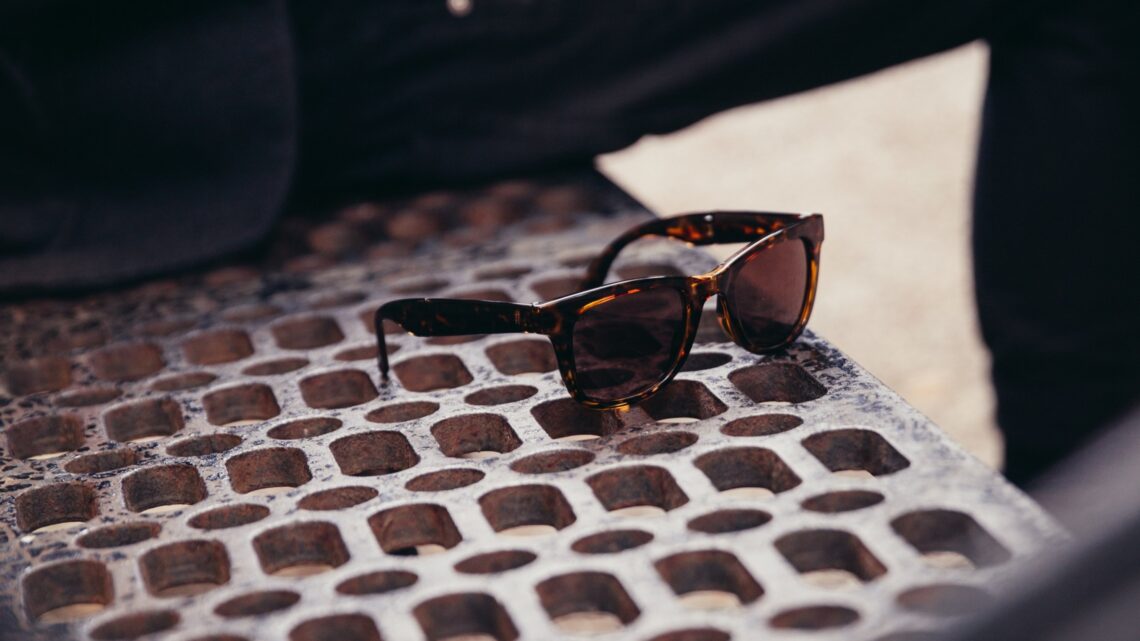A pair of wayfarer sunglasses kept on a bench beside a man.