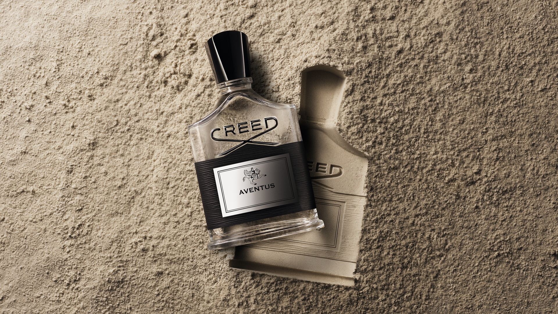 A Creed Aventus bottle on the sand, next to the impression of it.