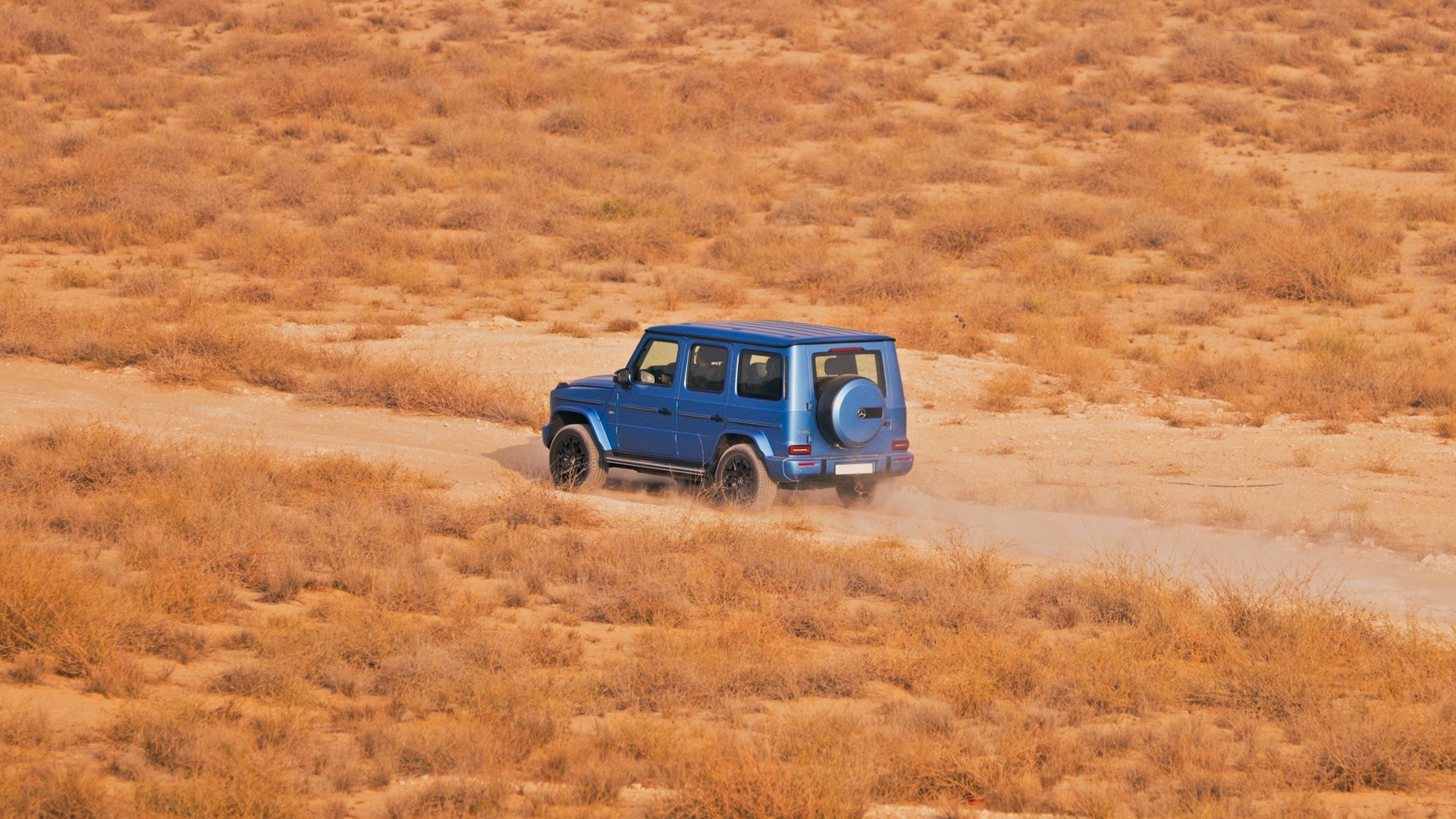 A dynamic shot of a blue Mercedes-Benz G 580, the Electric G-Wagen, taken from the rear-left side in an off-road terrain.